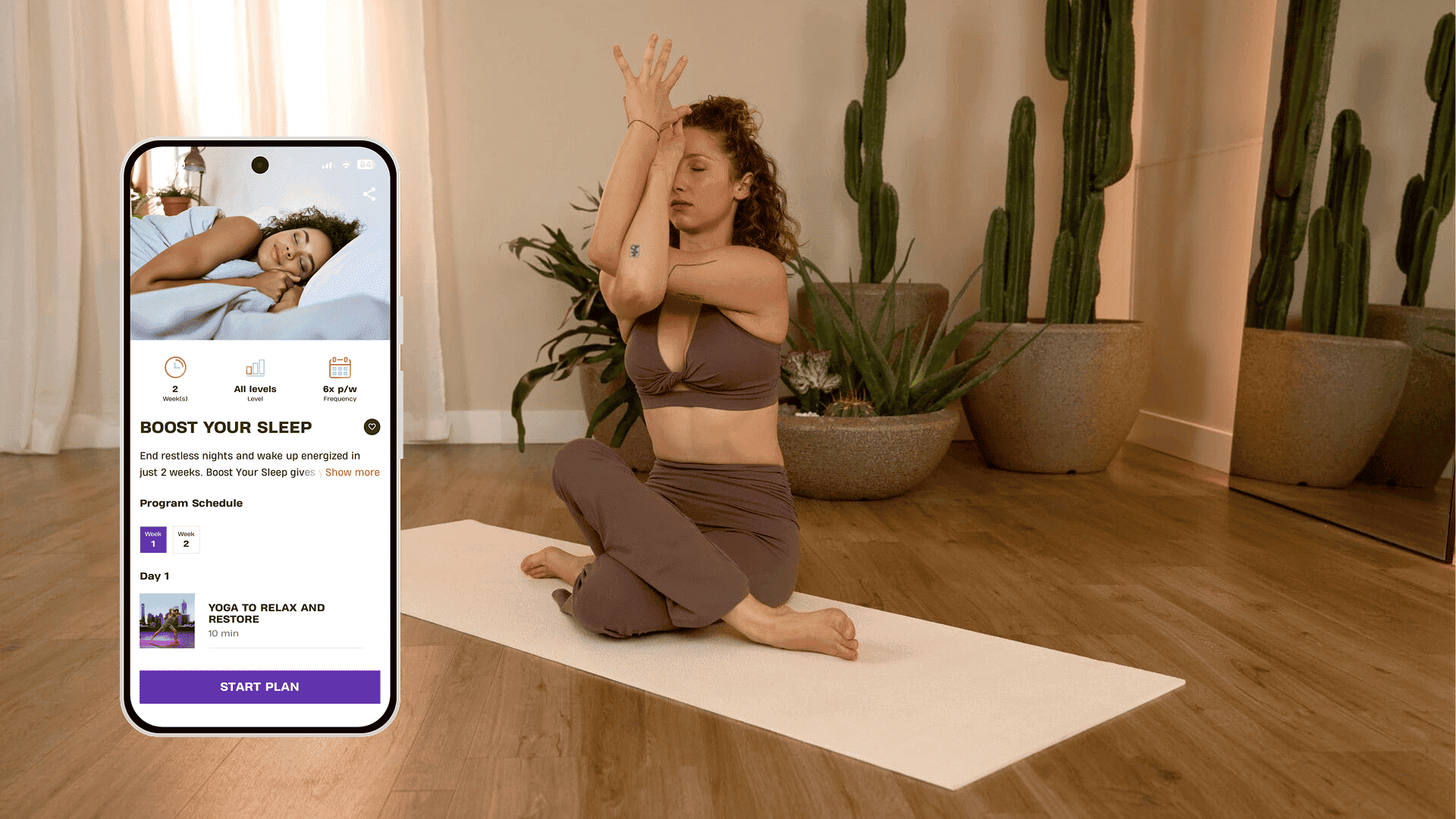 A woman practices yoga on a mat in a softly lit room surrounded by large potted plants, focusing on an arm and shoulder stretch while sitting cross-legged. Next to her, a smartphone screen shows the Basic-Fit app with the “Boost Your Sleep” program open, highlighting yoga sessions to relax and restore.