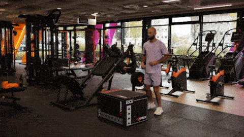 A person performs step-ups onto a sturdy plyo box in a Basic-Fit gym, stepping up and down with control.