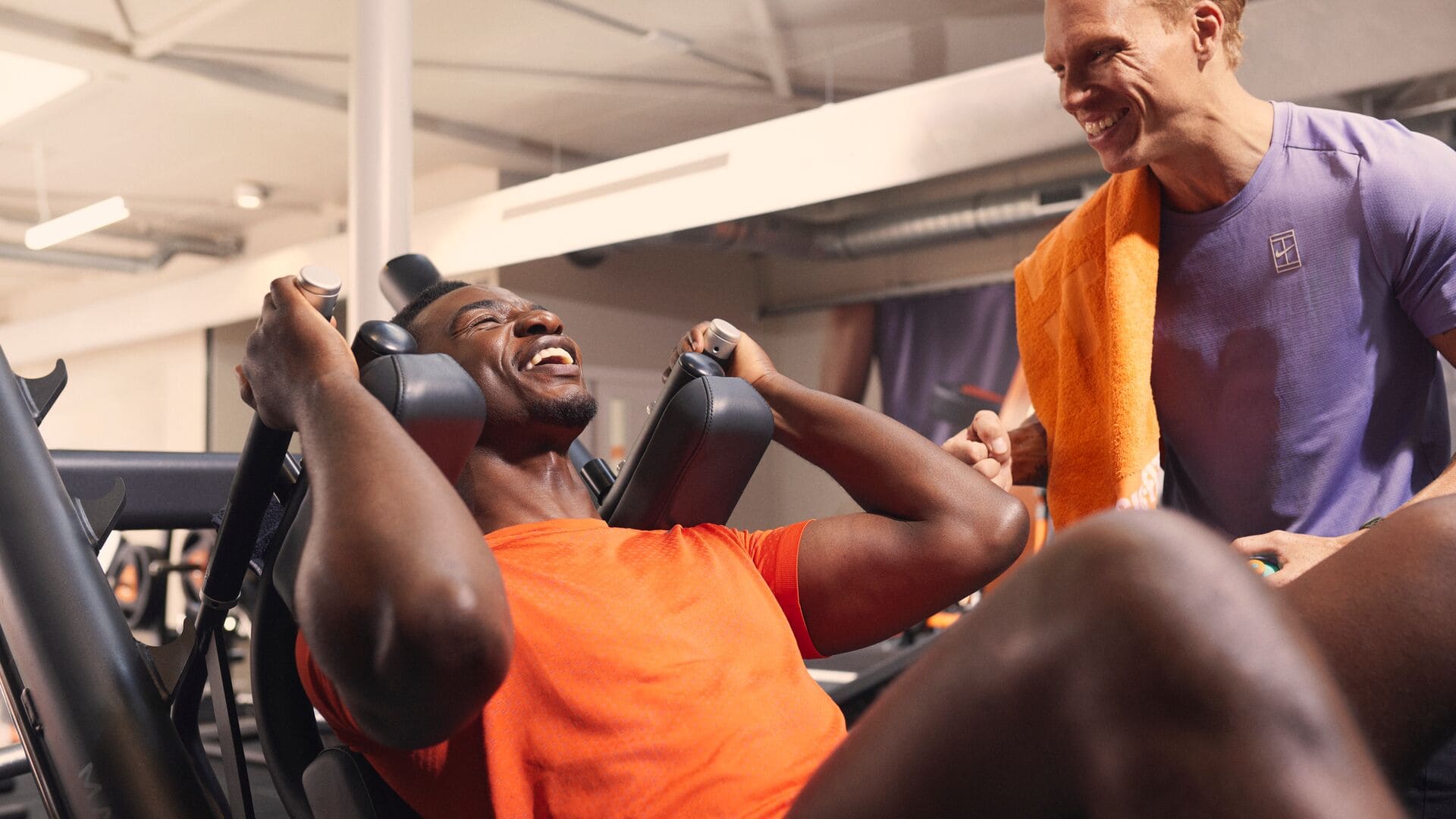 A man laughing while doing a leg press exercise, with a trainer beside him smiling and holding an orange towel. The atmosphere is friendly and energetic.