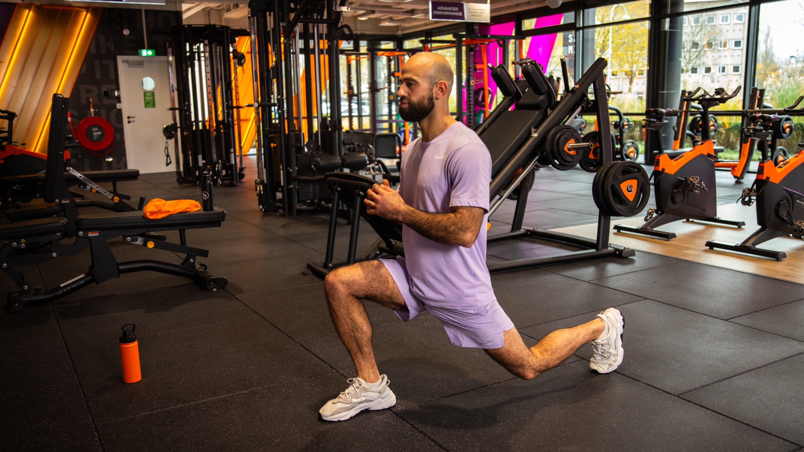 Person, die in einem Basic-Fit Club einen Ausfallschritt auf der Trainingsfläche ausführt, mit Kraftgeräten und Indoor-Bikes im Hintergrund.