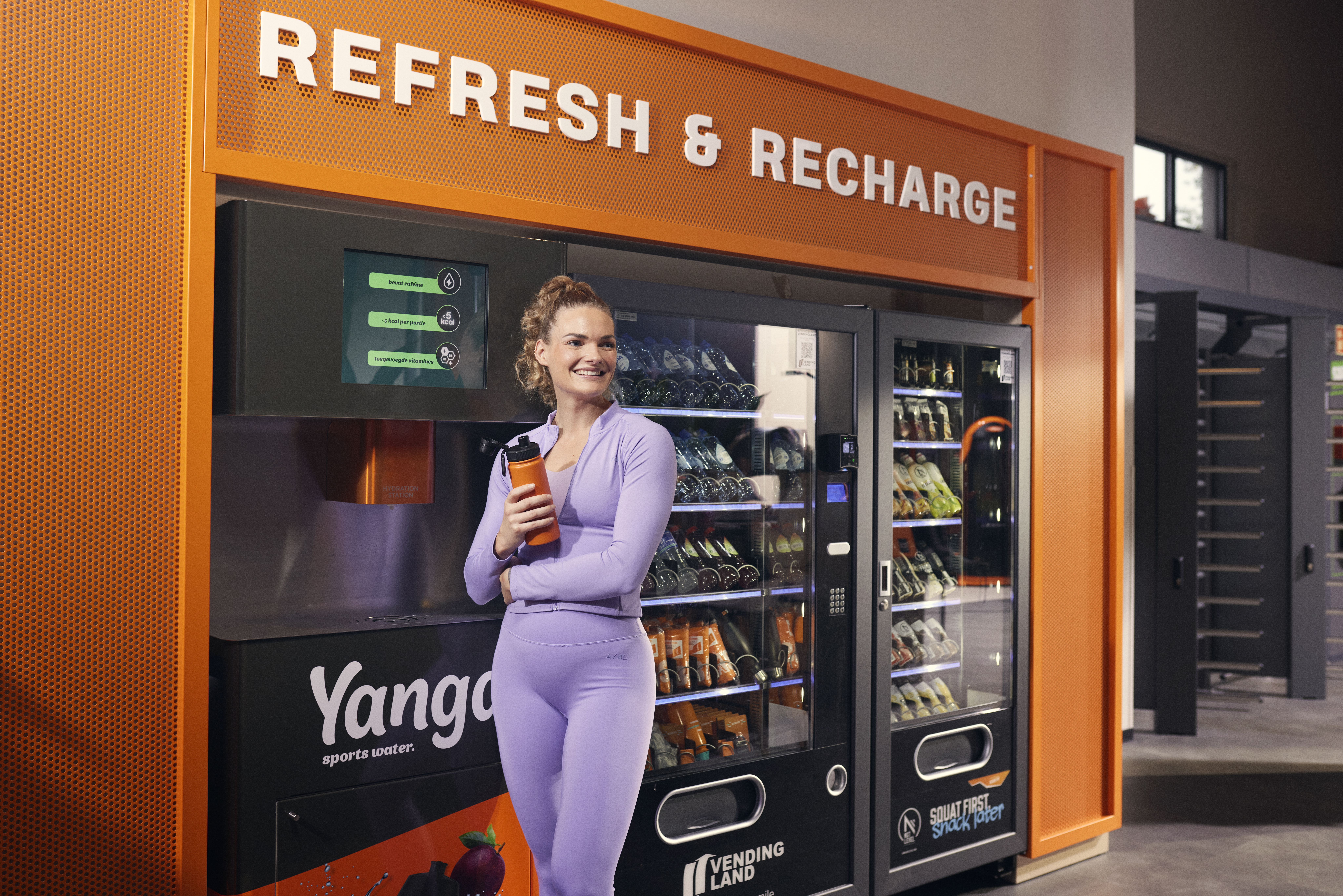 Una mujer con ropa deportiva morada sostiene una botella en la zona Refresh & Recharge con YANGA y máquinas expendedoras en Basic-Fit.