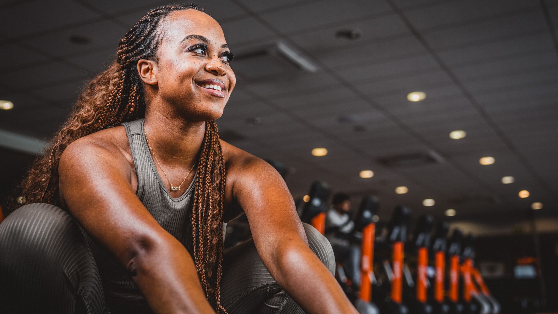 Femme en tenue de sport marron assise sur le sol de la salle de sport et souriant, avec des appareils de cardio et des accents orange en arrière-plan.