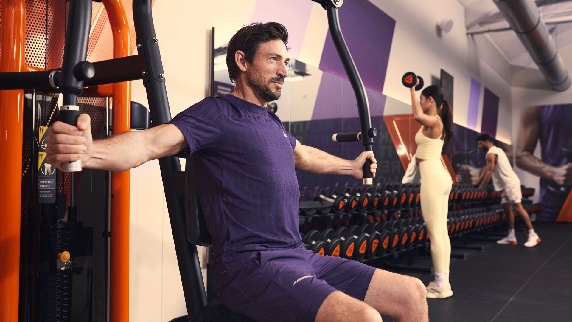 Man using a chest fly machine in the gym while other people lift weights in the background.