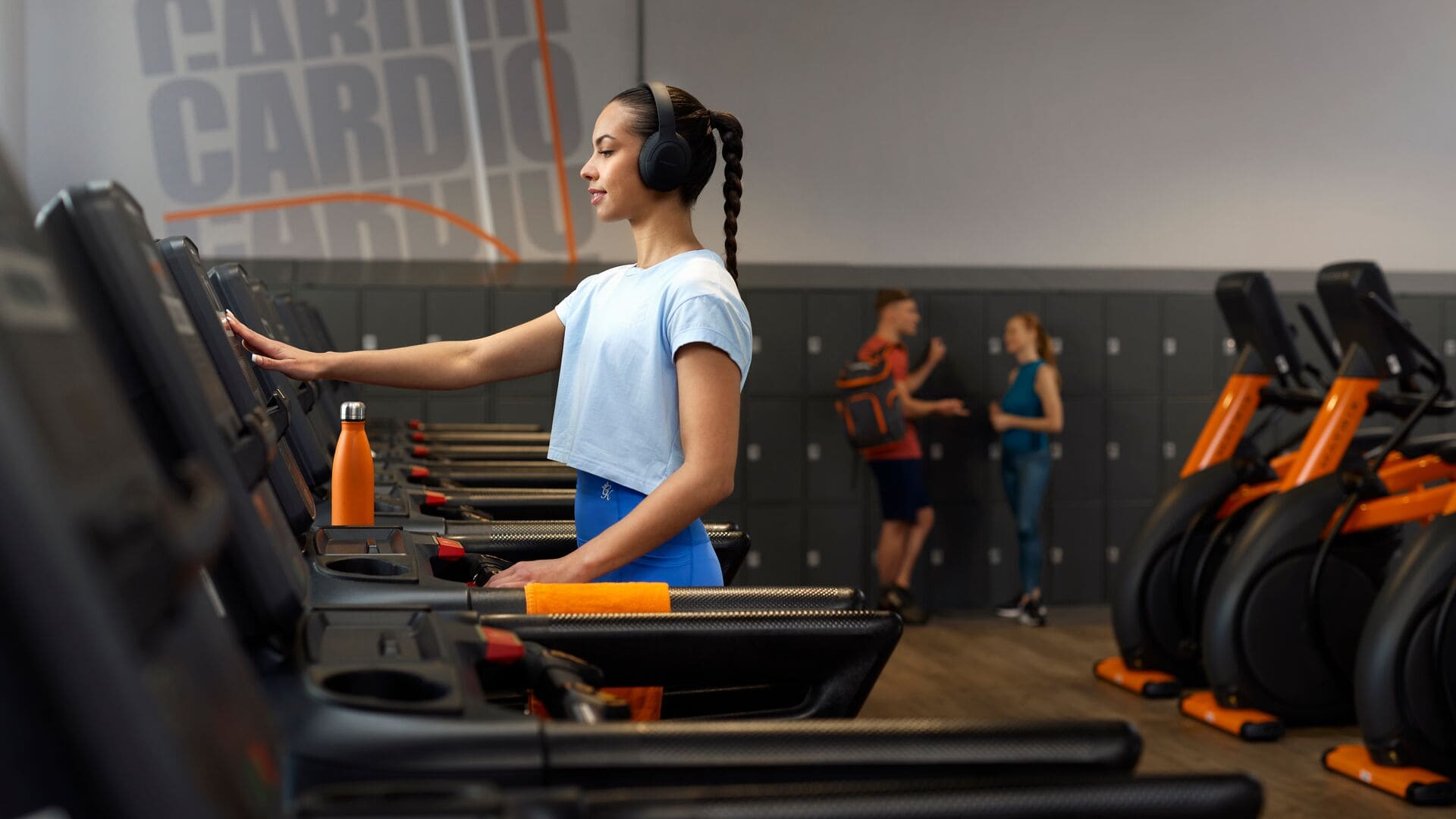 A woman in a light blue top and blue leggings uses a treadmill at Basic-Fit, wearing headphones, with an orange water bottle placed in front of her.