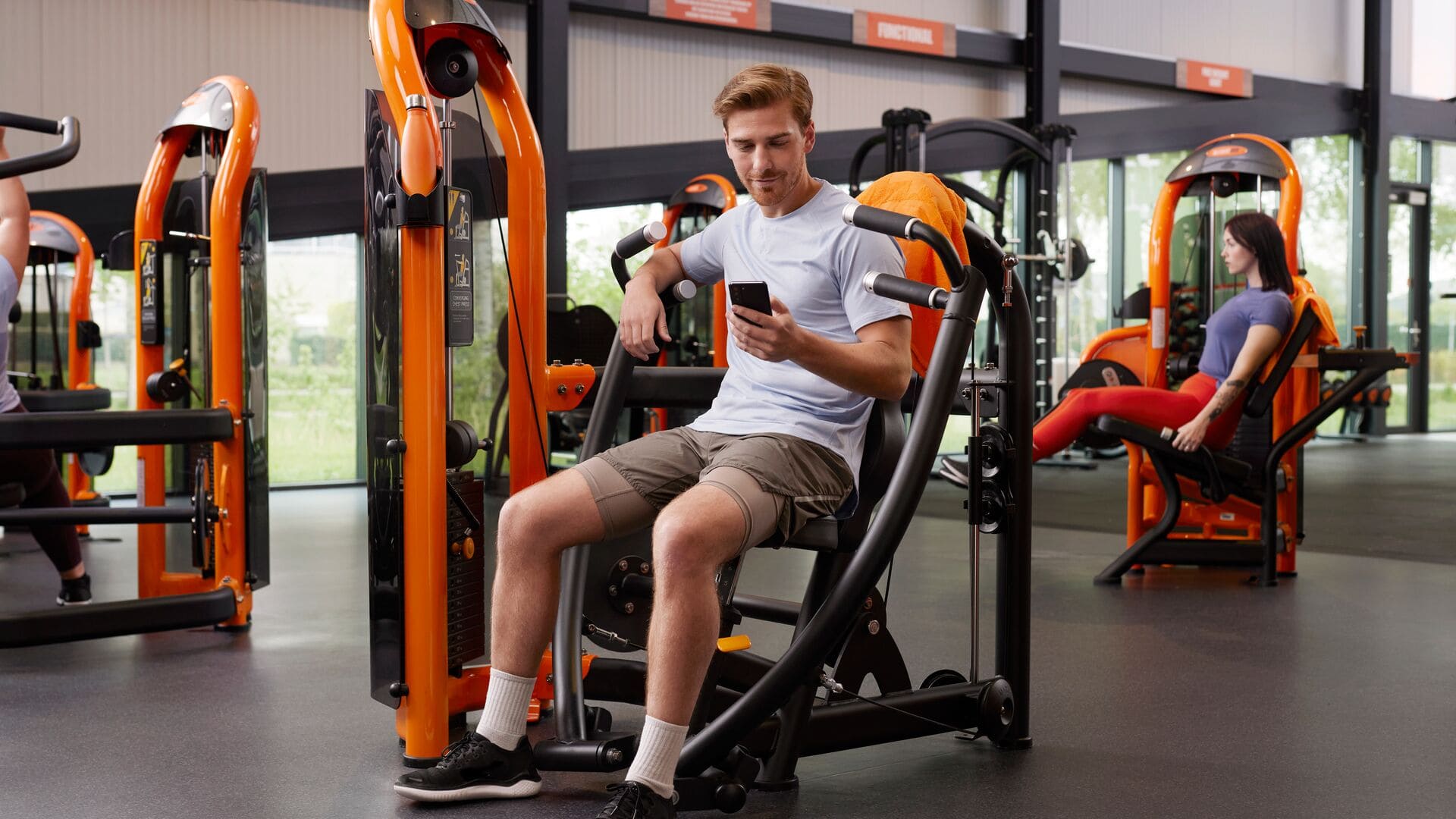 Un homme est assis sur une machine de presse à jambes dans une salle Basic-Fit et regarde son téléphone, tandis qu’une autre personne s’entraîne en arrière-plan.