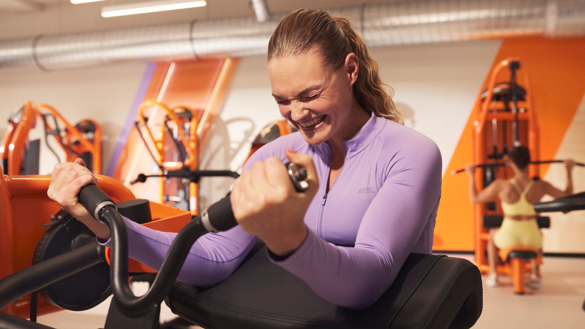 A woman performs a seated strength exercise on a machine in a Basic-Fit gym, focusing on her workout while smiling. Strength equipment and orange accents are visible in the background.