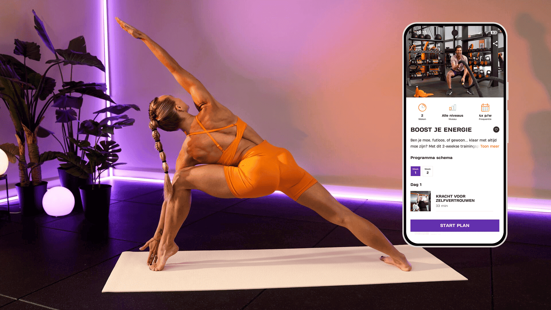 Een vrouw doet een diepe stretch in een yogahouding op een mat in een ruimte met paarse verlichting en tropische planten. Ze draagt feloranje sportkleding. Naast haar toont een smartphone het Basic-Fit-programma “Boost je energie” met trainingen die gericht zijn op kracht en zelfvertrouwen.
