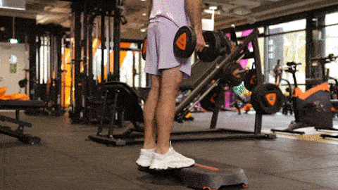 A person performs standing calf raises on a step platform in a Basic-Fit gym, lifting the heels up and down to work the calves.