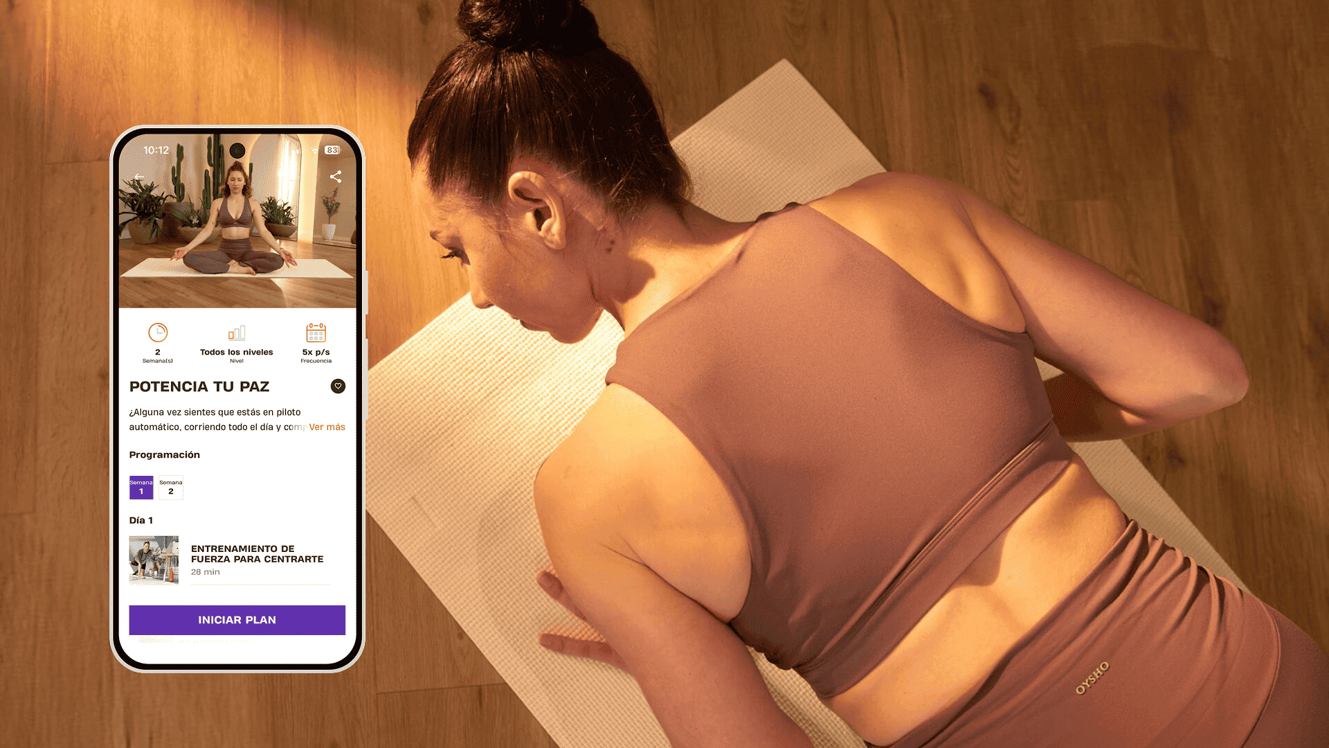 Mujer con ropa deportiva color malva acostada sobre una esterilla de yoga en una habitación con luz cálida y suelo de madera, junto a un teléfono con el programa de Basic-Fit “Potencia tu paz”.