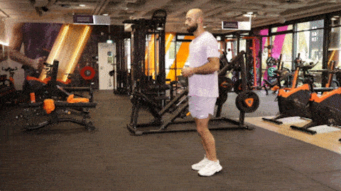 A person performs alternating forward lunges in a Basic-Fit gym, stepping forward and lowering into a lunge before switching legs.