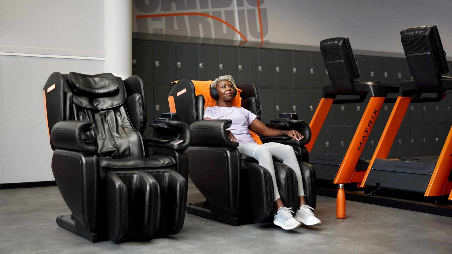 Une femme se détend dans un fauteuil de massage noir chez Basic-Fit, portant un casque audio, avec des tapis de course et des casiers en arrière-plan.