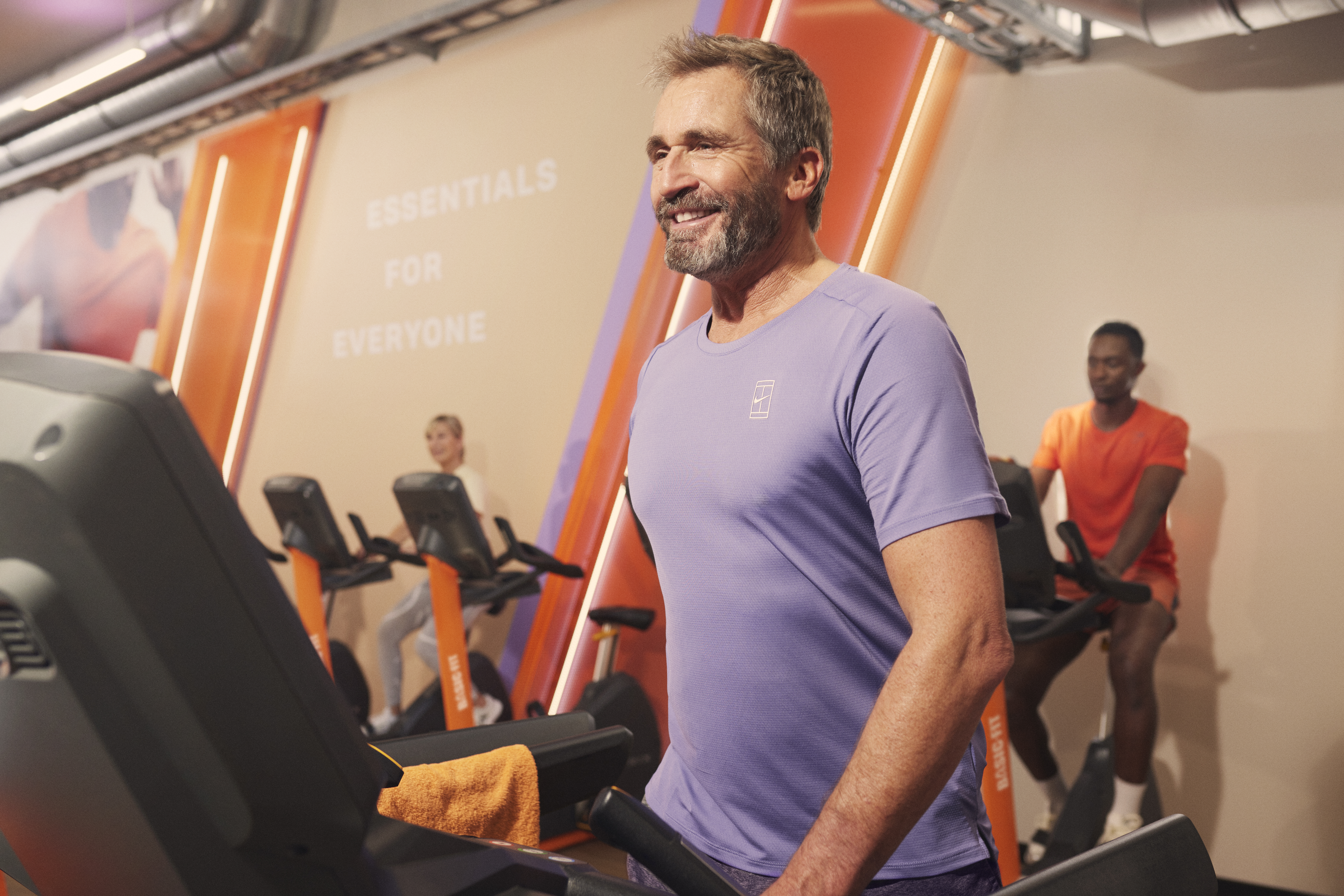 Un hombre con ropa deportiva morada sonríe entre las máquinas de cardio mientras otras personas entrenan al fondo en Basic-Fit.