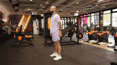 A person performs a bodyweight squat in a Basic-Fit gym, lowering into a controlled squat position and returning to standing on a training mat.