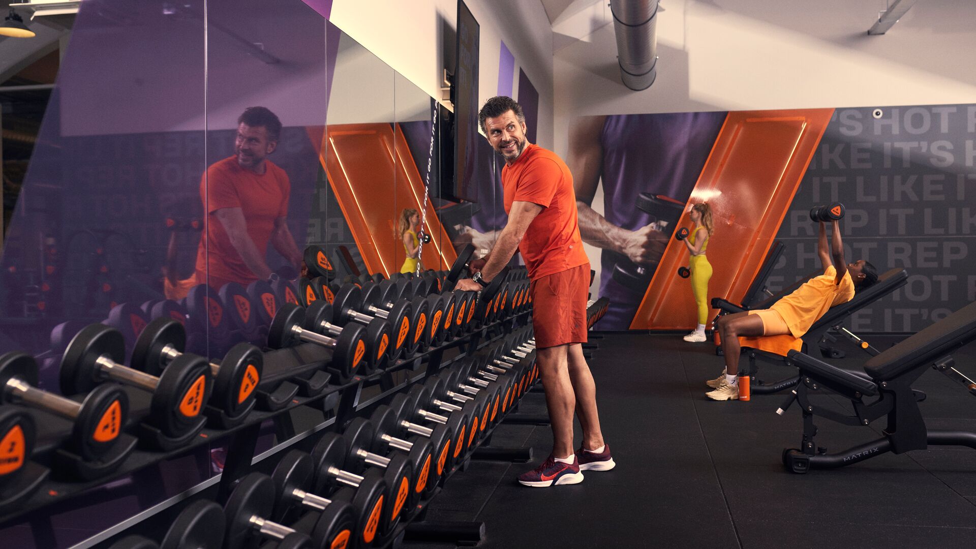 Hombre elige mancuernas en el gimnasio mientras otro miembro entrena en un banco al fondo