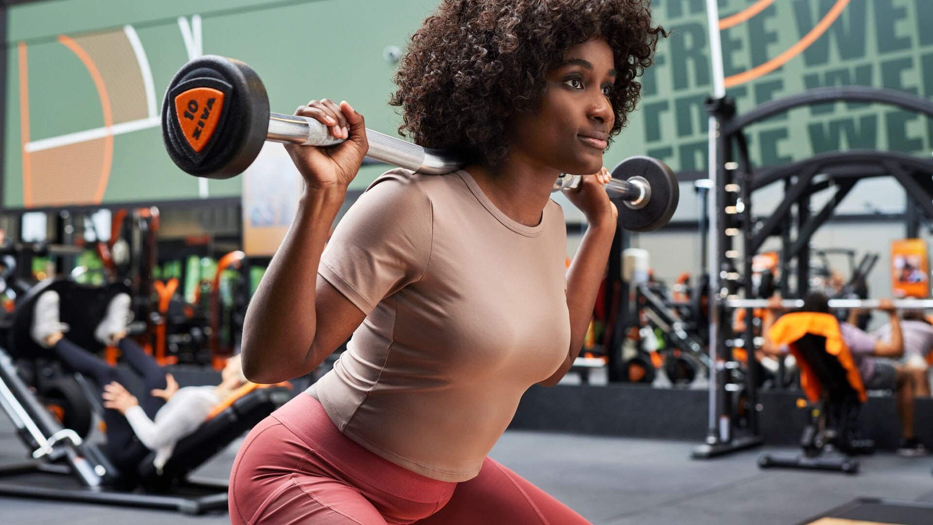 A woman performs a squat with a barbell across her shoulders in a Basic-Fit gym, focused and confident amid a dynamic workout environment with other members training in the background.        Ask ChatGPT