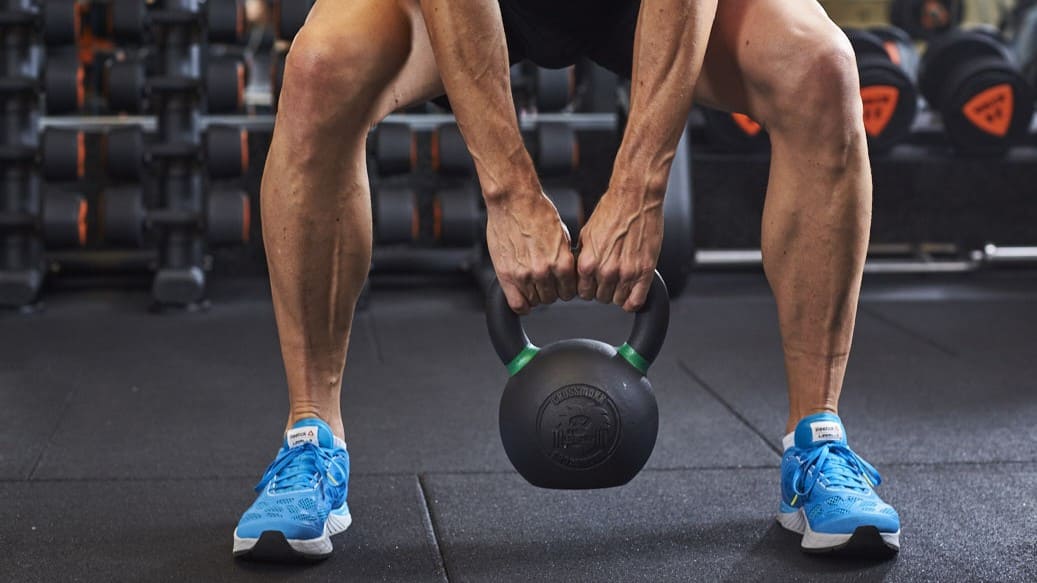 Person hebt eine Kettlebell in einem Basic-Fit-Studio, trägt blaue Sportschuhe und ist von Hanteln umgeben.