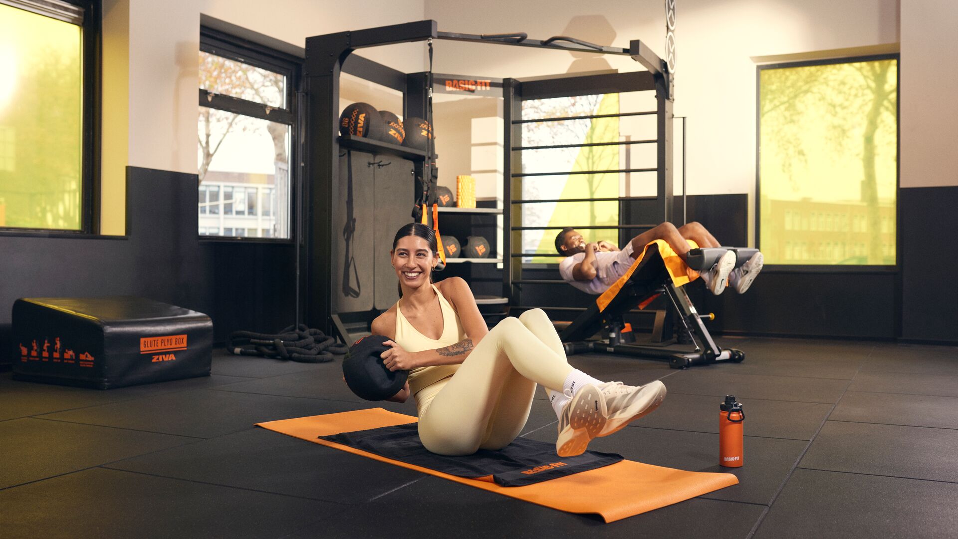 Eine Frau führt im Functional-Training-Bereich im Sitzen Russian Twists mit einem Medizinball auf einer orangefarbenen Trainingsmatte aus, während eine andere trainierende Person auf einer Negativbank trainiert.