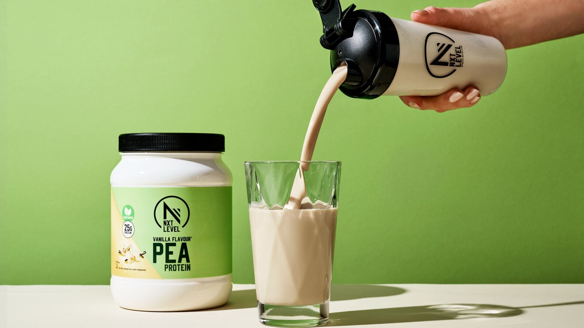 Hand gießt einen Vanille-Erbsenproteinshake in ein Glas neben einer NXT Level Protein-Dose vor grünem Hintergrund.