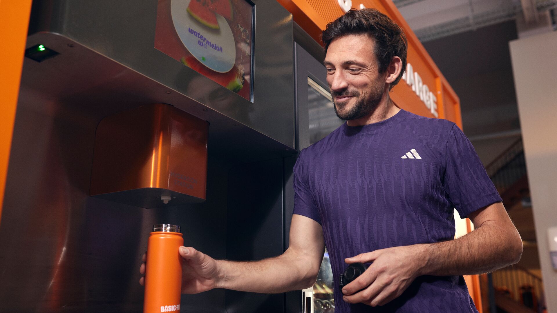 A man fills an orange Basic-Fit water bottle at a self service drink station inside a Basic-Fit gym.