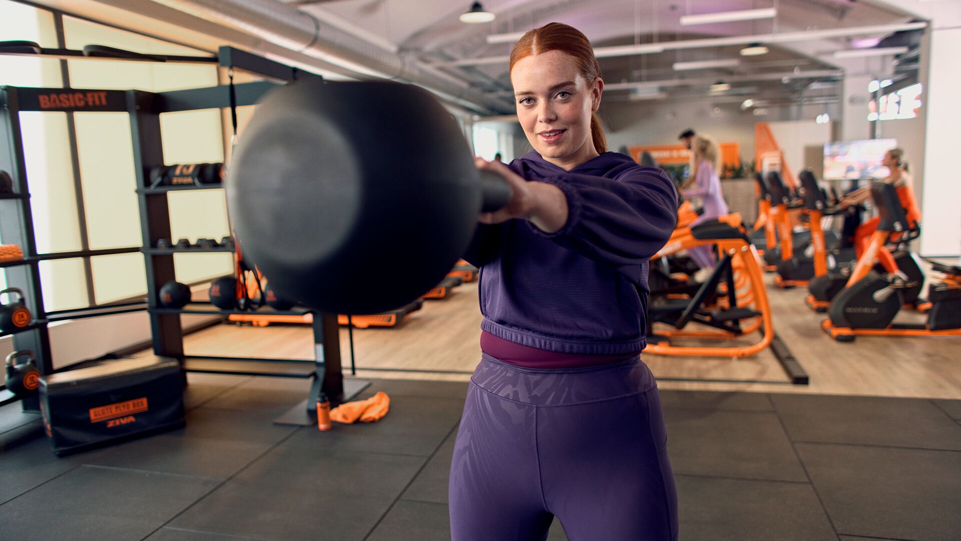 Woman performing a kettlebell swing in the functional training area of a Basic-Fit gym, with cardio machines in the background.