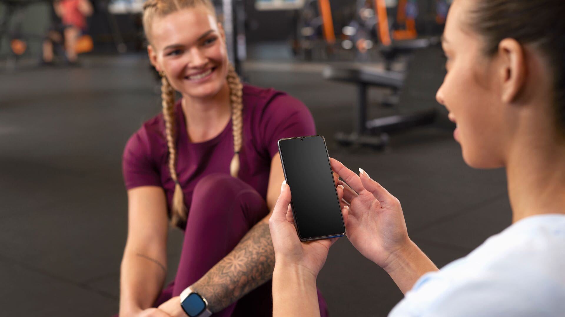 Two women at the gym sit on the floor after training; one holds up a smartphone while the other smiles, both in sportswear.