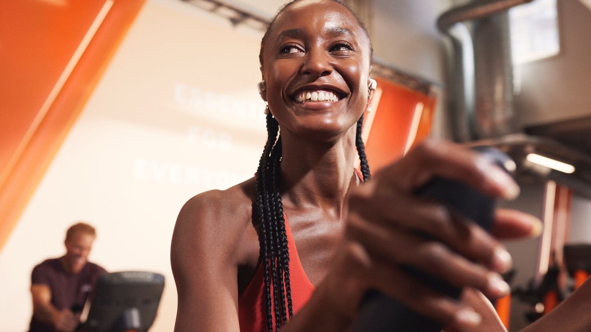 A woman smiling while using a cardio machine in a brightly lit gym with orange accents. She is wearing wireless earphones and a red sports outfit, with other people training in the background.