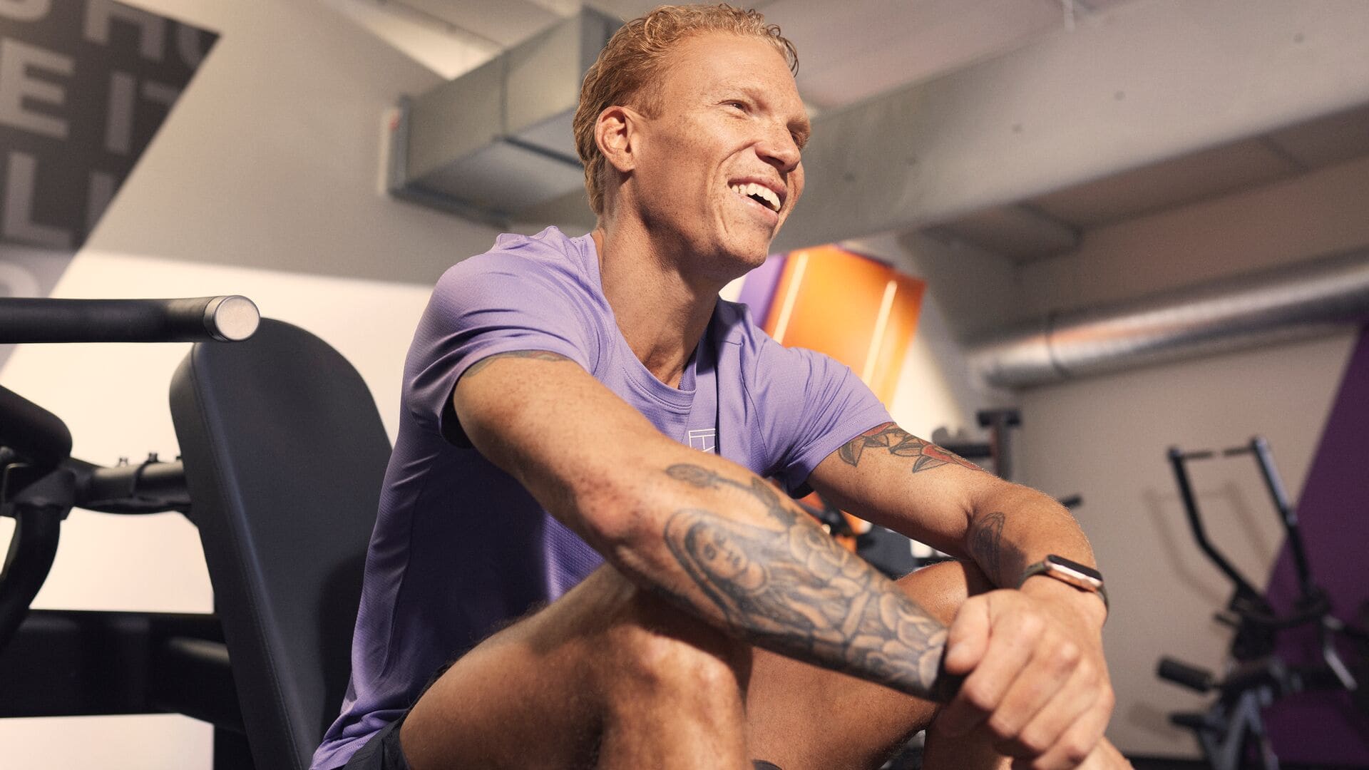A man with tattoos sitting on a gym bench, resting after a workout and smiling. The gym background shows equipment and orange accents.