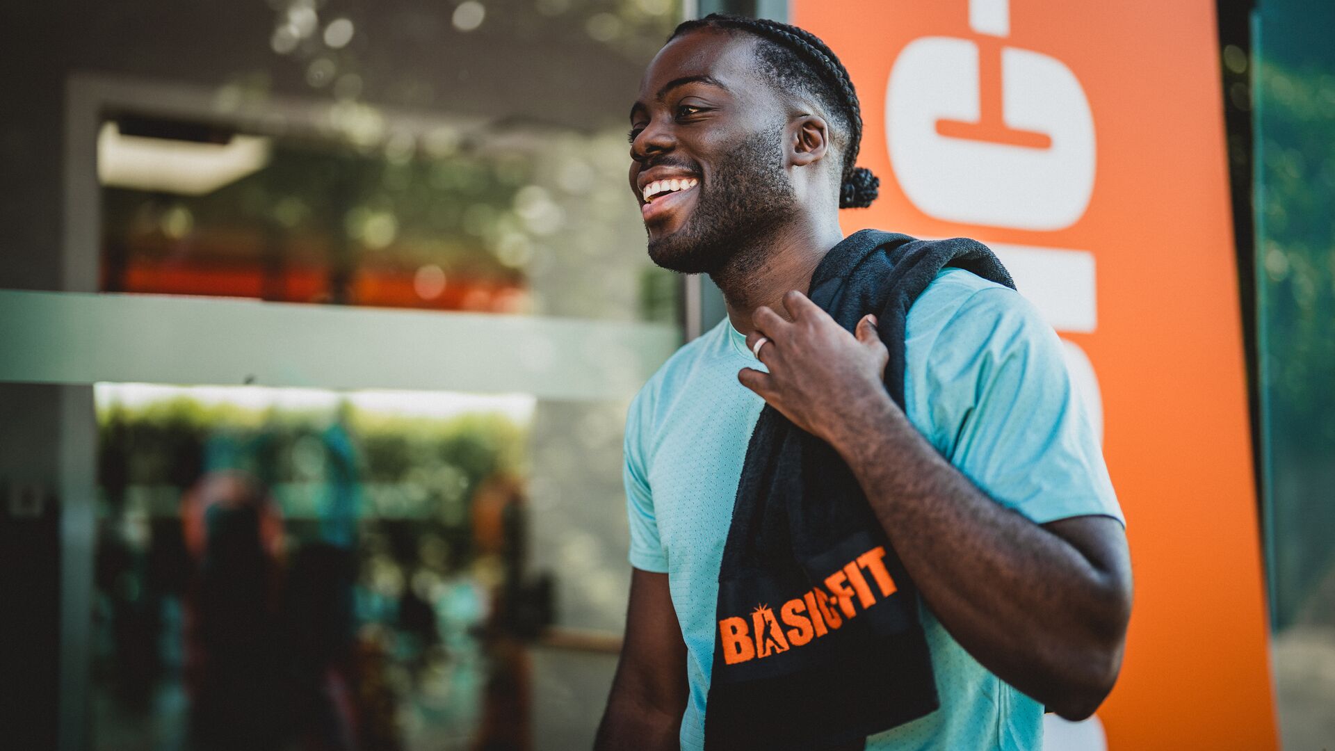 Homme souriant en T-shirt bleu clair devant un club Basic-Fit, portant une serviette noire avec le logo orange Basic-Fit sur l’épaule.