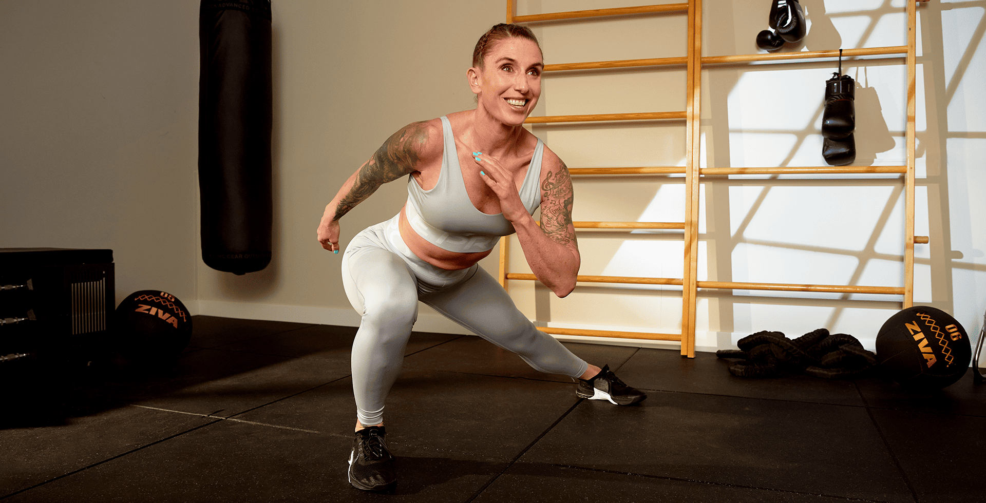 A woman in light workout gear smiles while doing a dynamic lateral lunge in a Basic-Fit training zone, surrounded by functional fitness equipment like a punching bag, medicine balls and battle ropes.