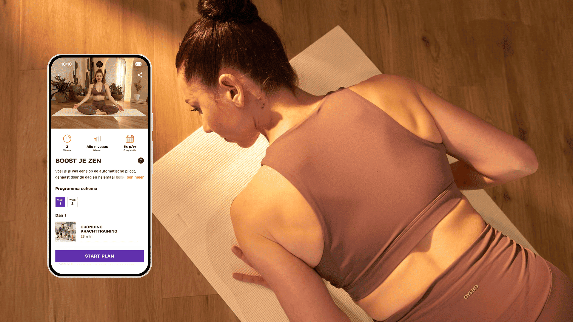 Een vrouw ligt op een yogamat in een warm verlichte ruimte, klaar voor een rustige workout. Ze draagt mauvekleurige sportkleding en richt zich op haar ademhaling. Naast haar toont een smartphone het Basic-Fit-programma “Boost je zen”, met sessies om balans en innerlijke kracht te herstellen.