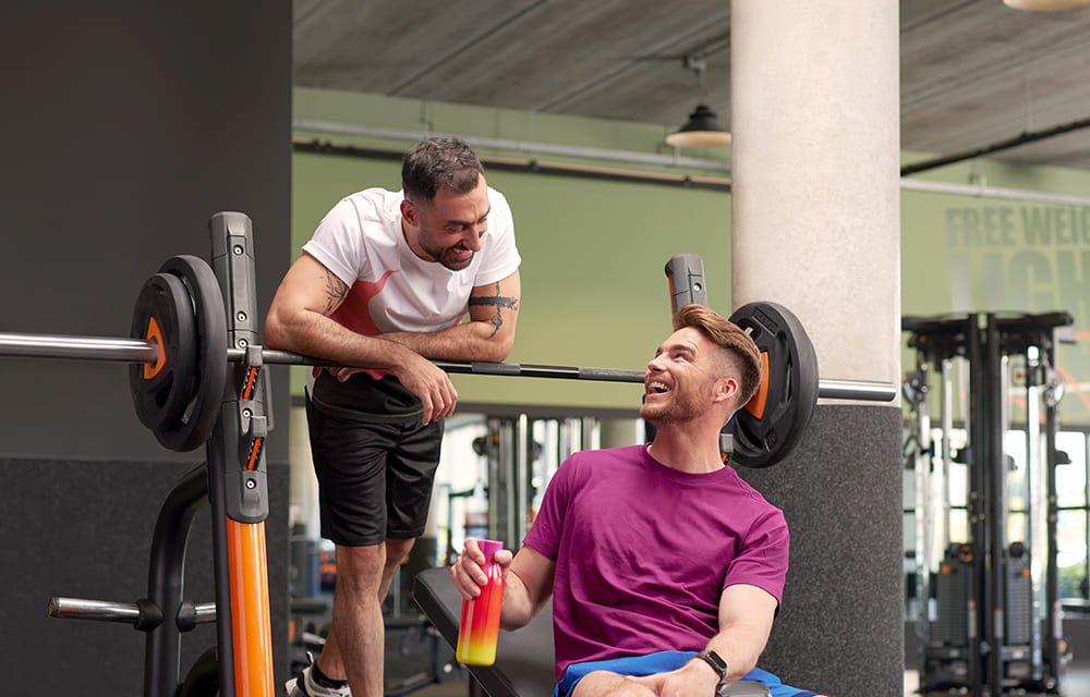 Twee mannen zijn in een Basic-Fit sportschool aan het praten en lachen bij een halter op een rek. De één staat in een wit-rood shirt en de ander zit in een paars shirt met een shakebeker in zijn hand.