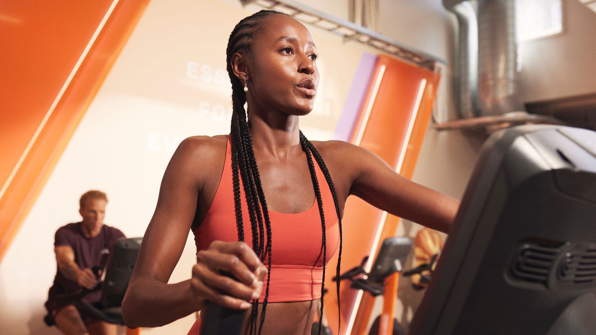A woman in a red sports outfit working out on a cardio machine in a gym with orange lighting. She looks determined while exercising.