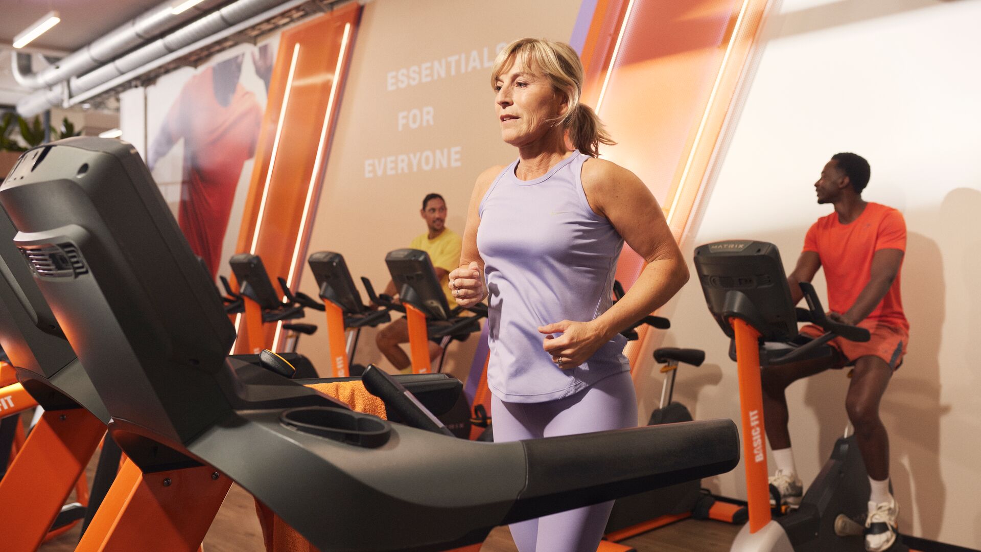 Eine Frau in fliederfarbener Sportkleidung joggt auf einem Laufband in einem Basic-Fit-Studio, während andere Trainierende im Hintergrund Fahrradergometer zwischen orangefarbenen Fitnessgeräten nutzen.