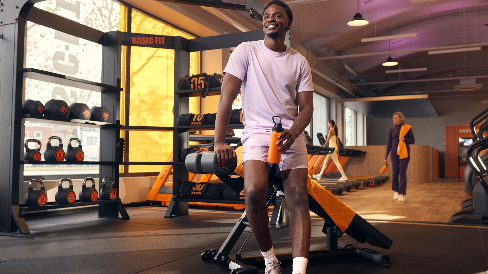 Personne assise sur un banc de musculation tenant une gourde dans une salle de sport, avec des kettlebells et des appareils de cardio visibles autour.
