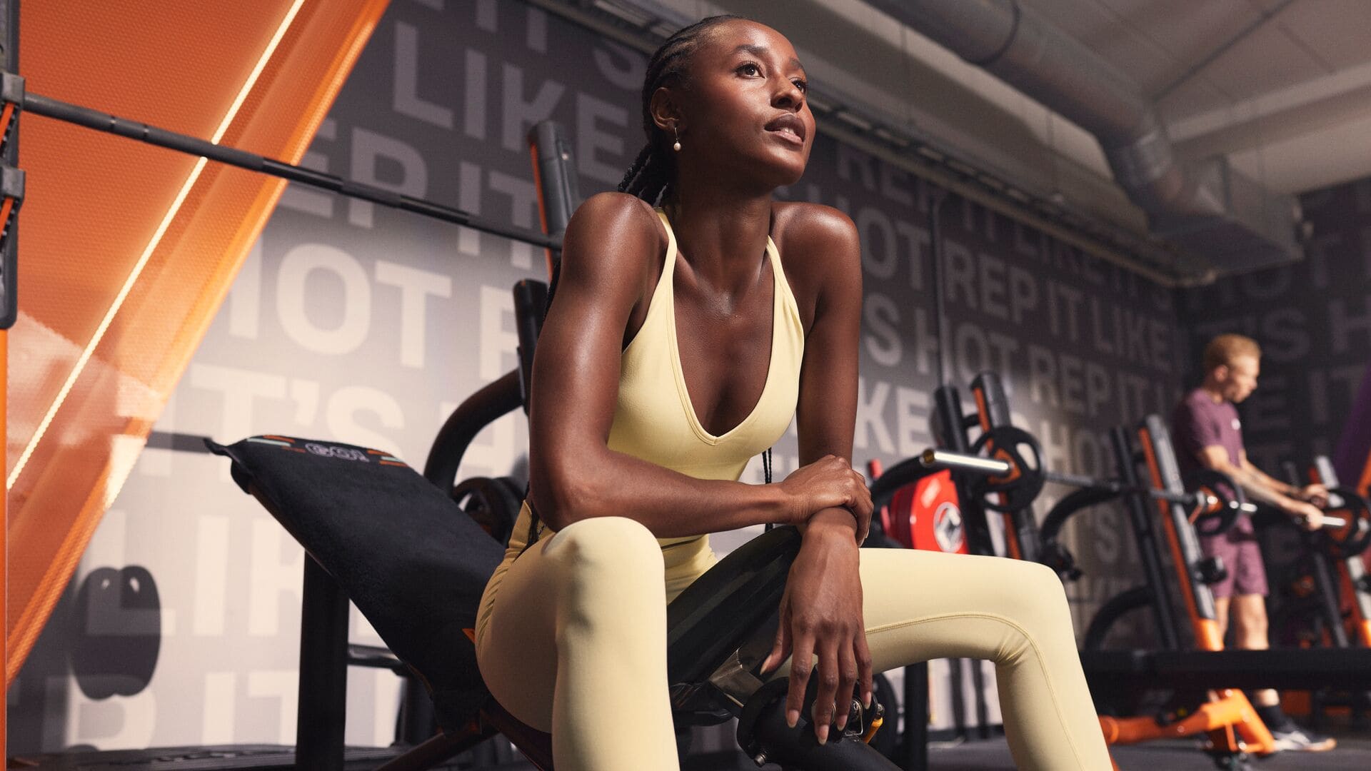 A woman in a yellow sports outfit sitting on a workout bench, resting after exercise. She looks focused while other people train in the background.