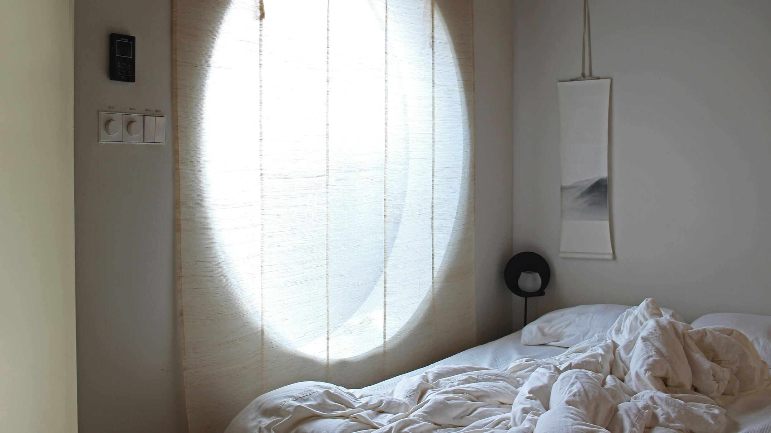 Soft morning light shines through sheer curtains into a peaceful bedroom with an unmade bed and neutral tones.