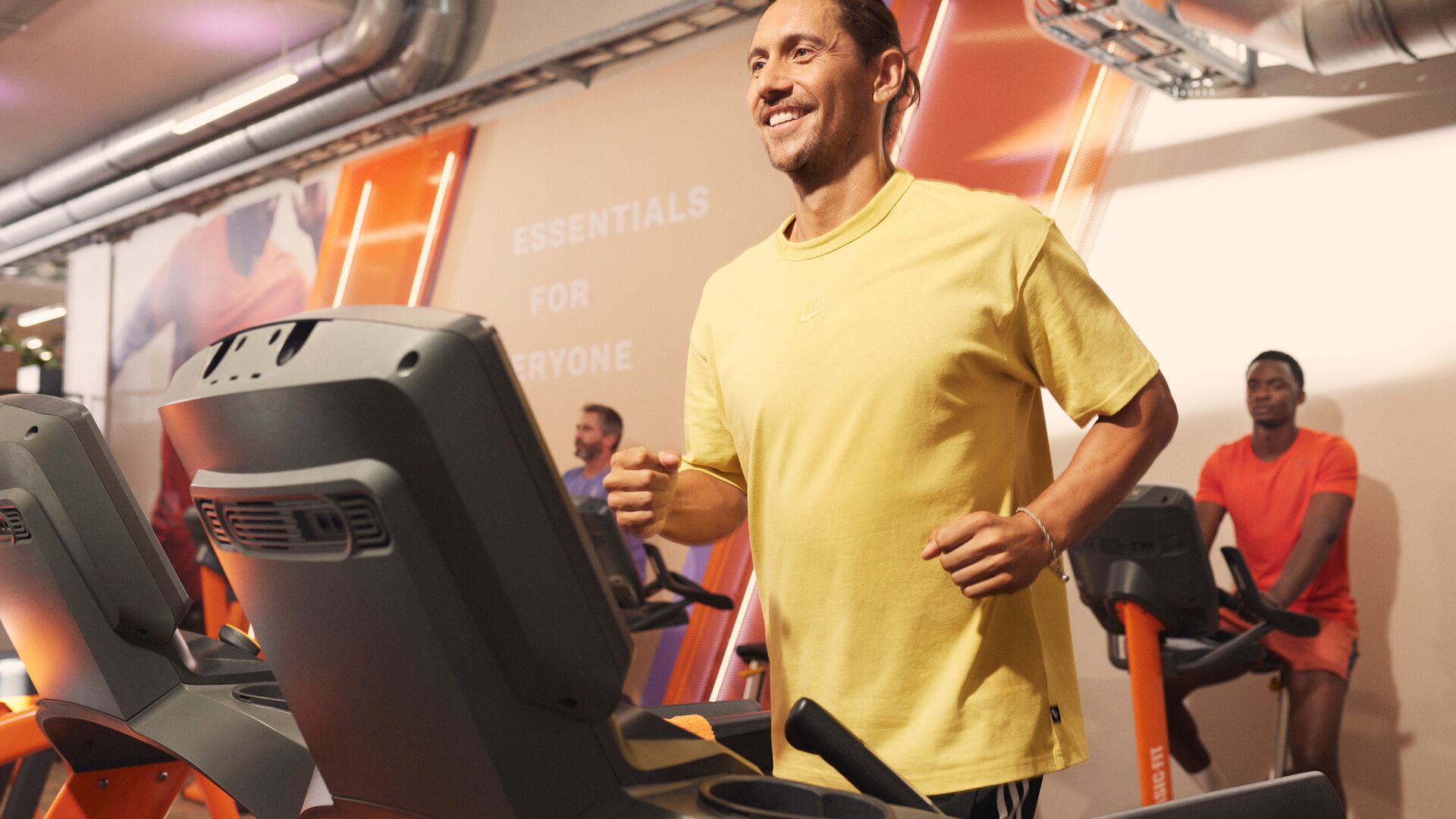 A man runs on a treadmill in a Basic-Fit gym, wearing a yellow shirt, with other members training on cardio machines behind him in a brightly lit space with orange details.