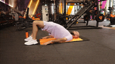 A person performs a glute bridge on a mat in a Basic-Fit gym, lifting the hips off the floor and lowering them with control.