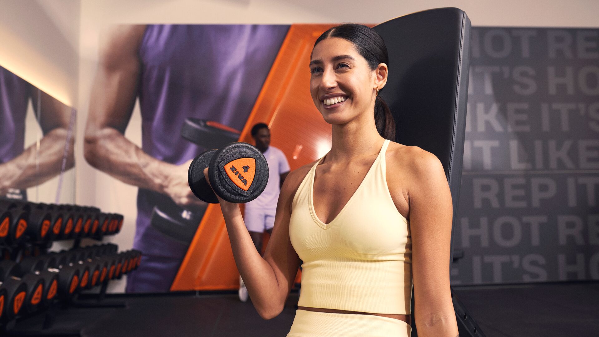 Person smiling while performing a dumbbell bicep curl in a gym, standing next to a rack of dumbbells with strength equipment in the background.