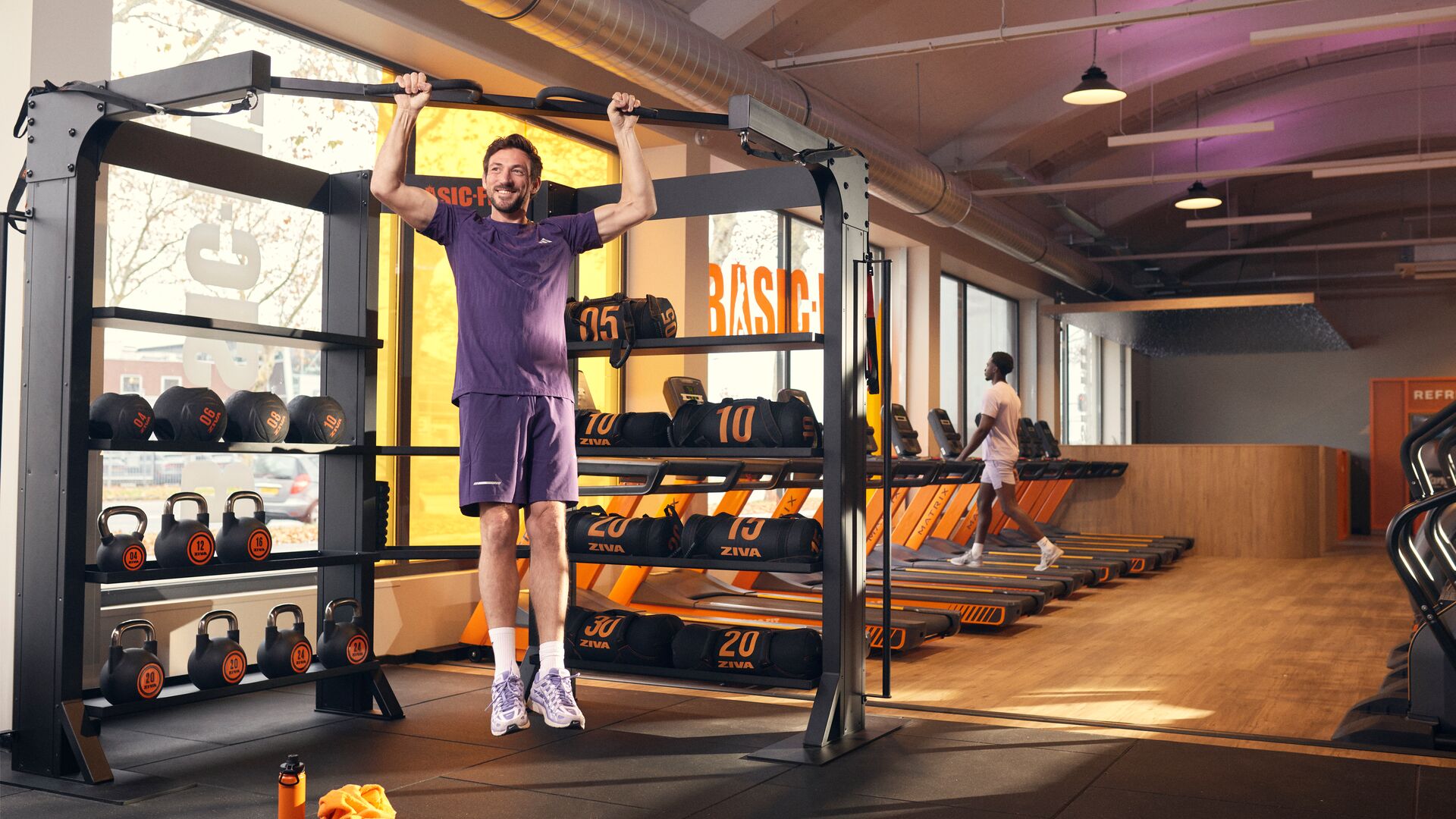 Person doing a pull-up on a bar in a gym, positioned near free weights and treadmills, with daylight coming through large windows.