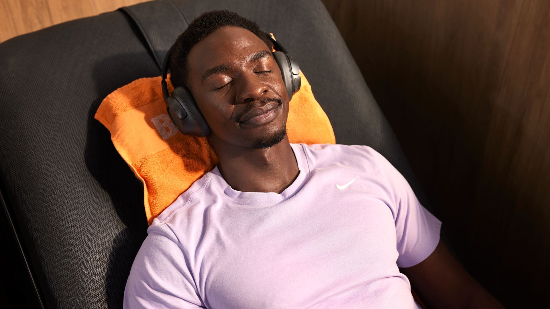 A man in a light purple sports shirt relaxes with headphones on, lying back with eyes closed on an orange towel in a Basic-Fit massage chair.