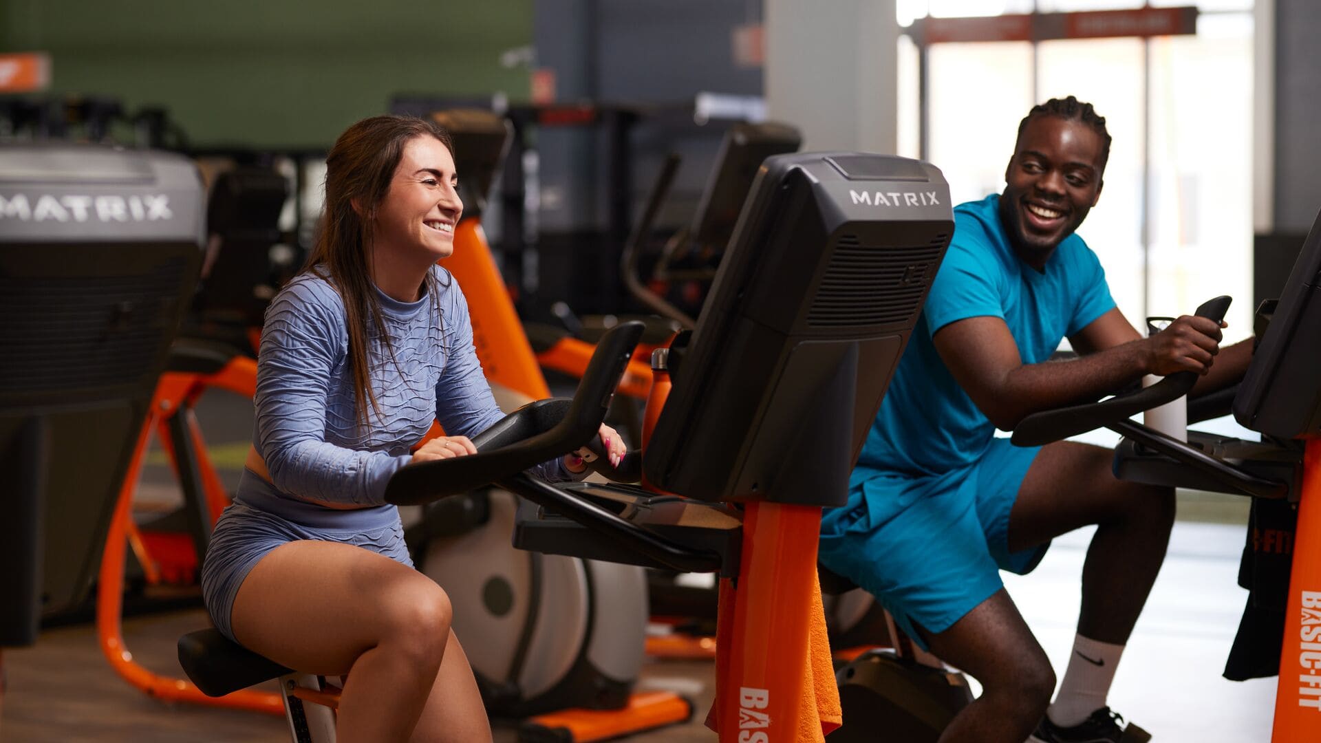 Een vrouw in een lichtblauwe sportset en een man in een turquoise outfit zitten op hometrainers in een Basic-Fit sportschool, lachend terwijl ze met elkaar praten tijdens hun cardiotraining.