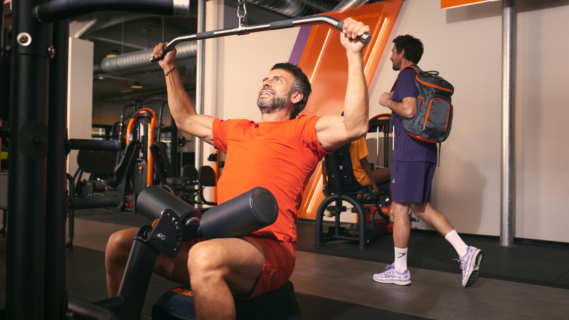 Man performing a lat pulldown exercise on a machine in a gym, with other members and equipment visible in the background.
