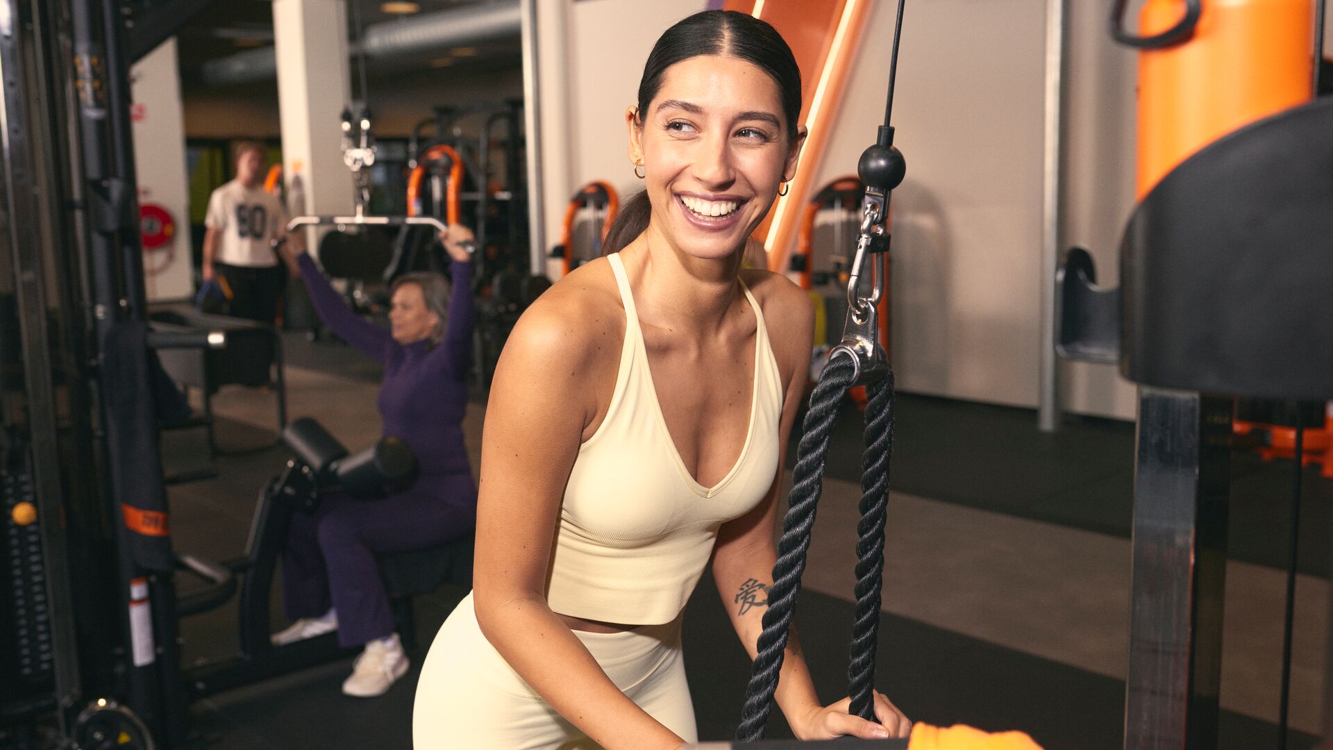 A woman smiles while using a cable machine in a Basic-Fit gym, holding a rope attachment during a strength exercise. Other members train in the background.