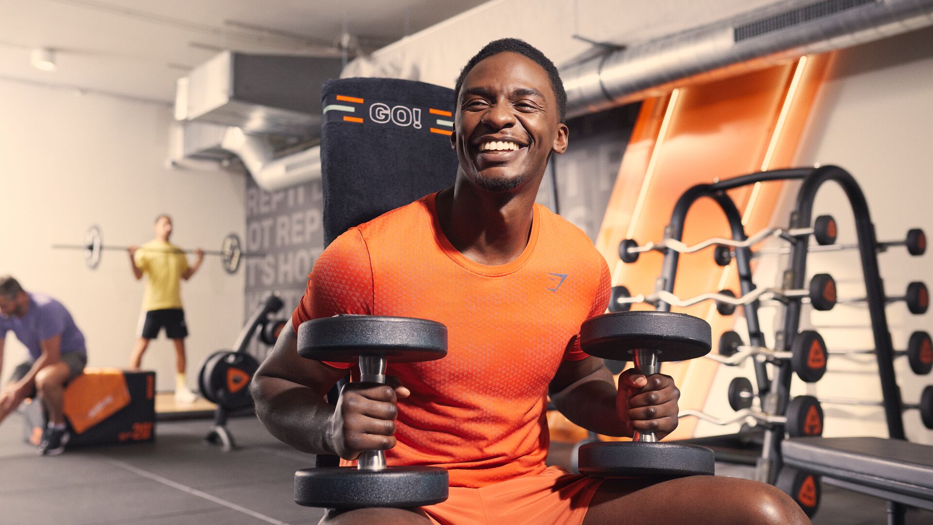 A man smiles while seated on a bench in a Basic-Fit gym, holding two dumbbells during a strength workout. Other members train in the background among strength equipment and orange accents.