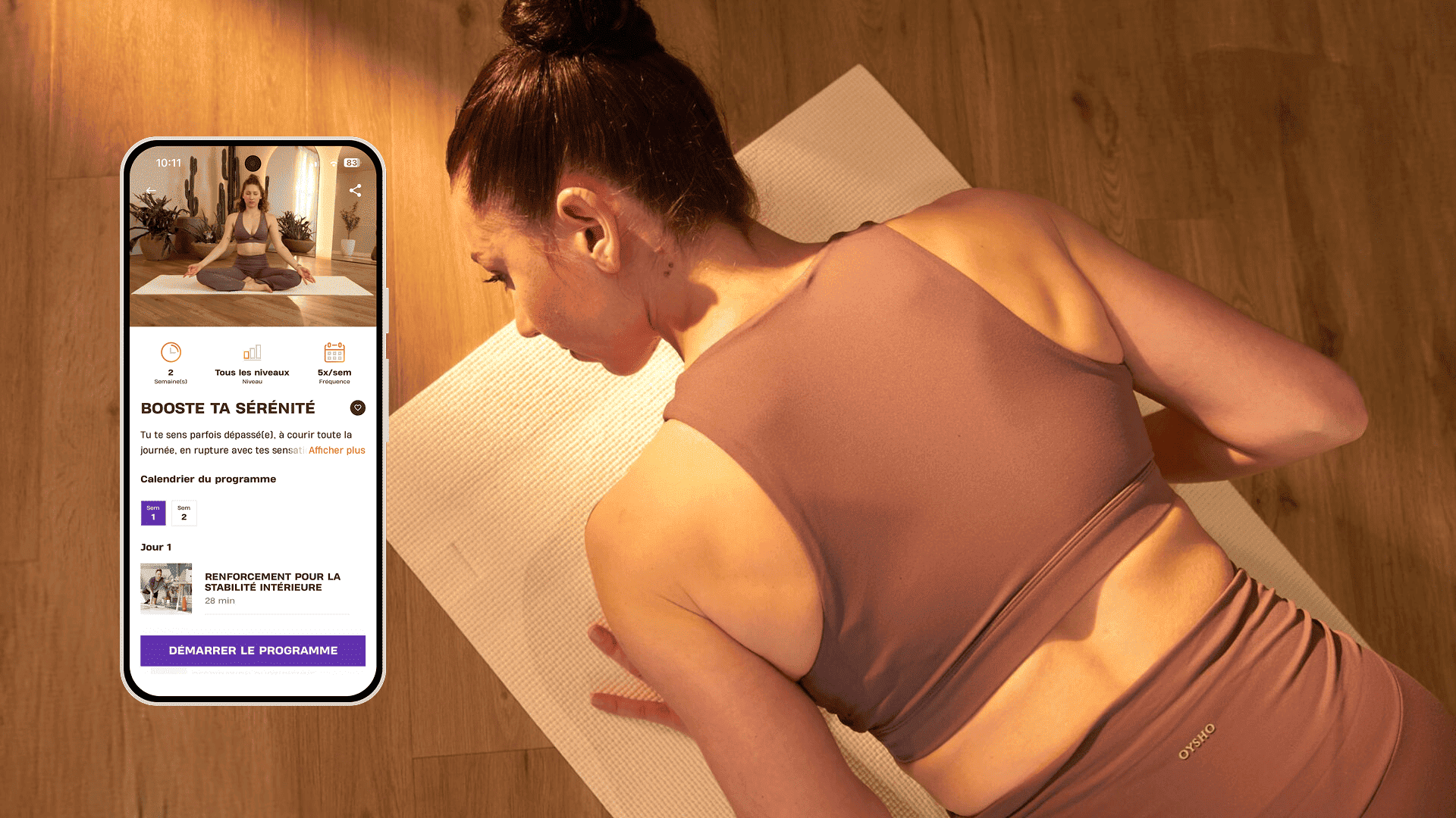 Une femme est allongée sur un tapis de yoga dans une pièce doucement éclairée, prête pour une séance apaisante. Elle porte une tenue de sport couleur mauve et se concentre sur sa respiration. À côté d’elle, un smartphone affiche le programme Basic-Fit “Booste ta sérénité”, proposant des séances pour retrouver équilibre et force intérieure.